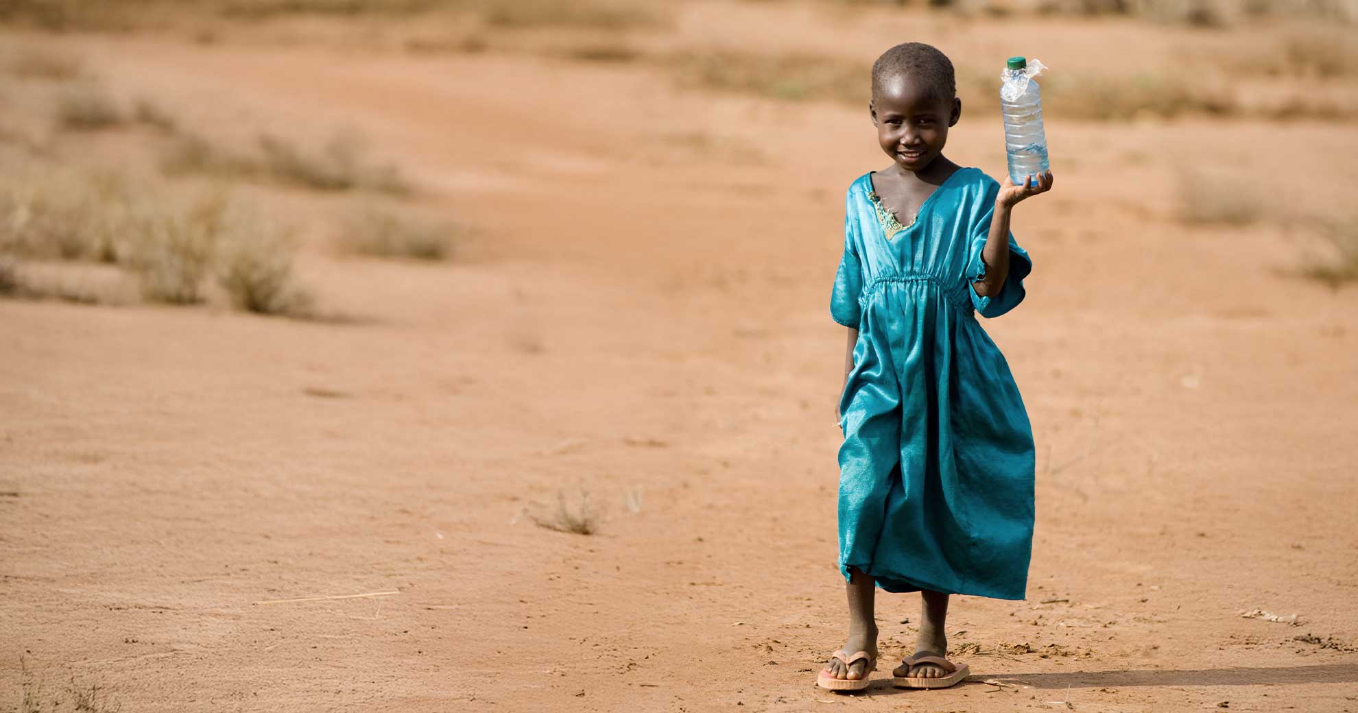 Kind mit Wasserflasche als Symbol fuer Trinkwasserprojekte Ein Kind in blauem Kleid haelt eine Wasserflasche und laechelt, Symbol fuer den Zugang zu sauberem Trinkwasser