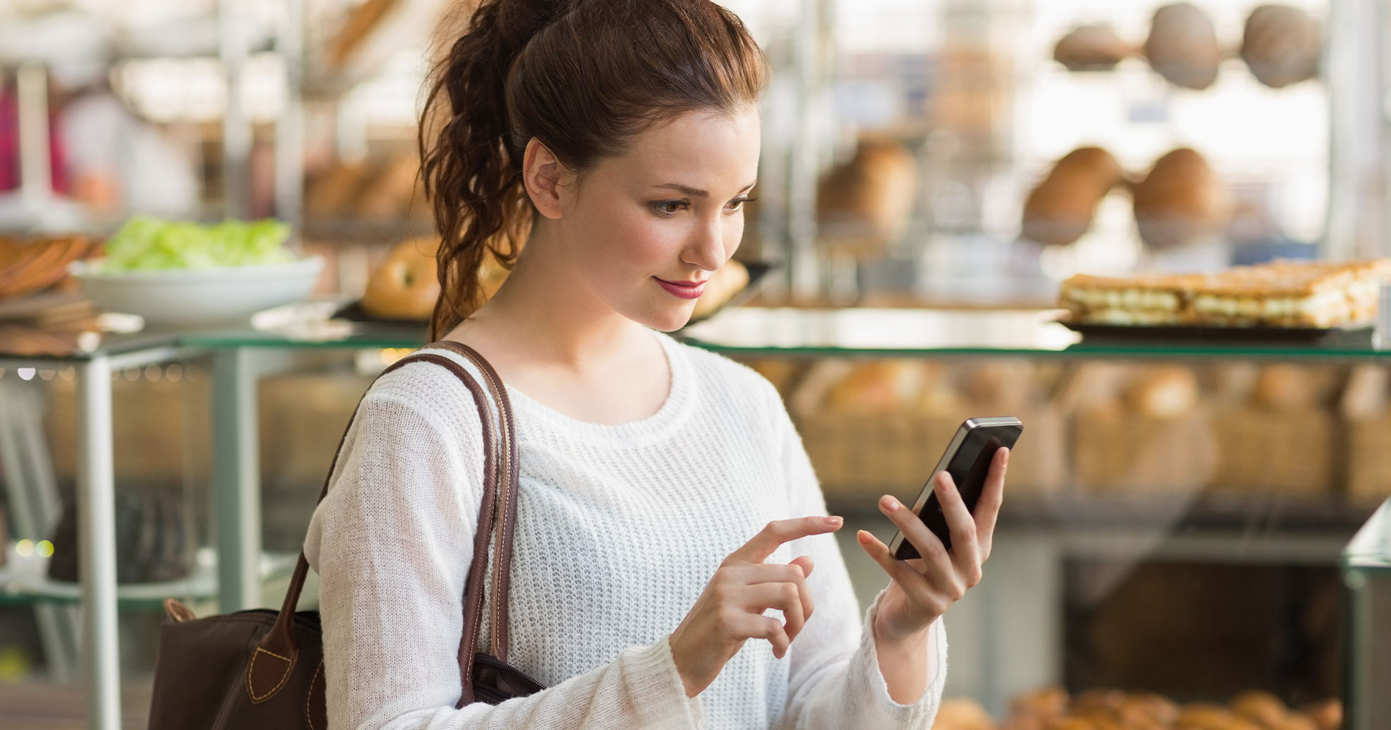 Junge Frau mit Smartphone in Bäckerei beim Reservieren von Lebensmitteln über mobile App, Gebäck und Brötchen im Hintergrund
